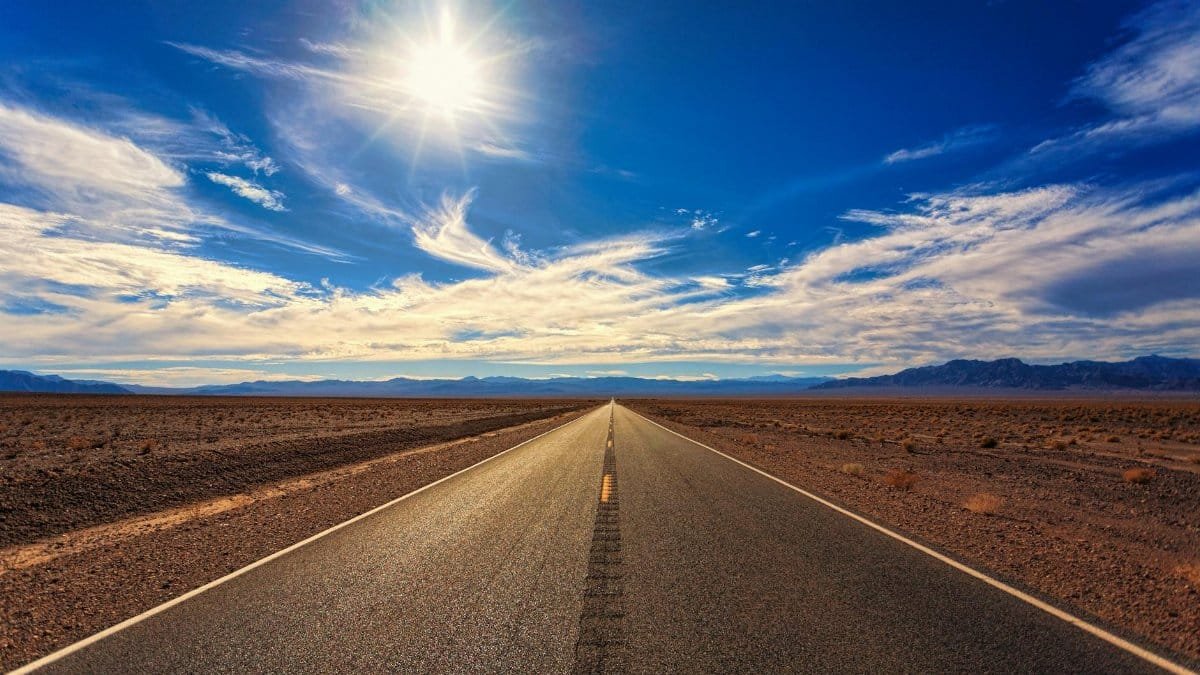 A long, straight road stretches across a dry desert landscape under a bright blue sky and shining sun.