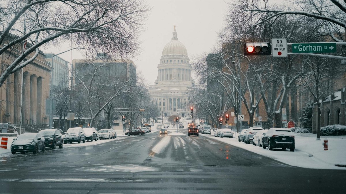 A serene winter view of the Wisconsin State Capitol under snowfall with snow-covered streets and cars.