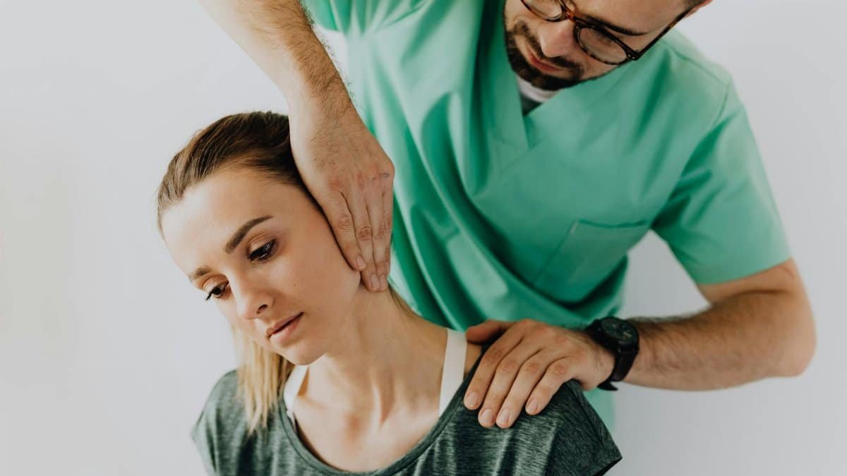 Confident doctor wearing uniform and eyeglasses gently doing therapeutic massage on calm female patients neck and stretching stiff neck muscles