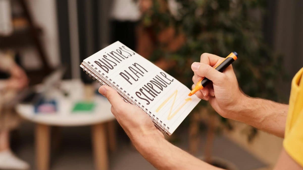 Close-up of a hand writing in a notebook with 'Business Plan Schedule' indoors.