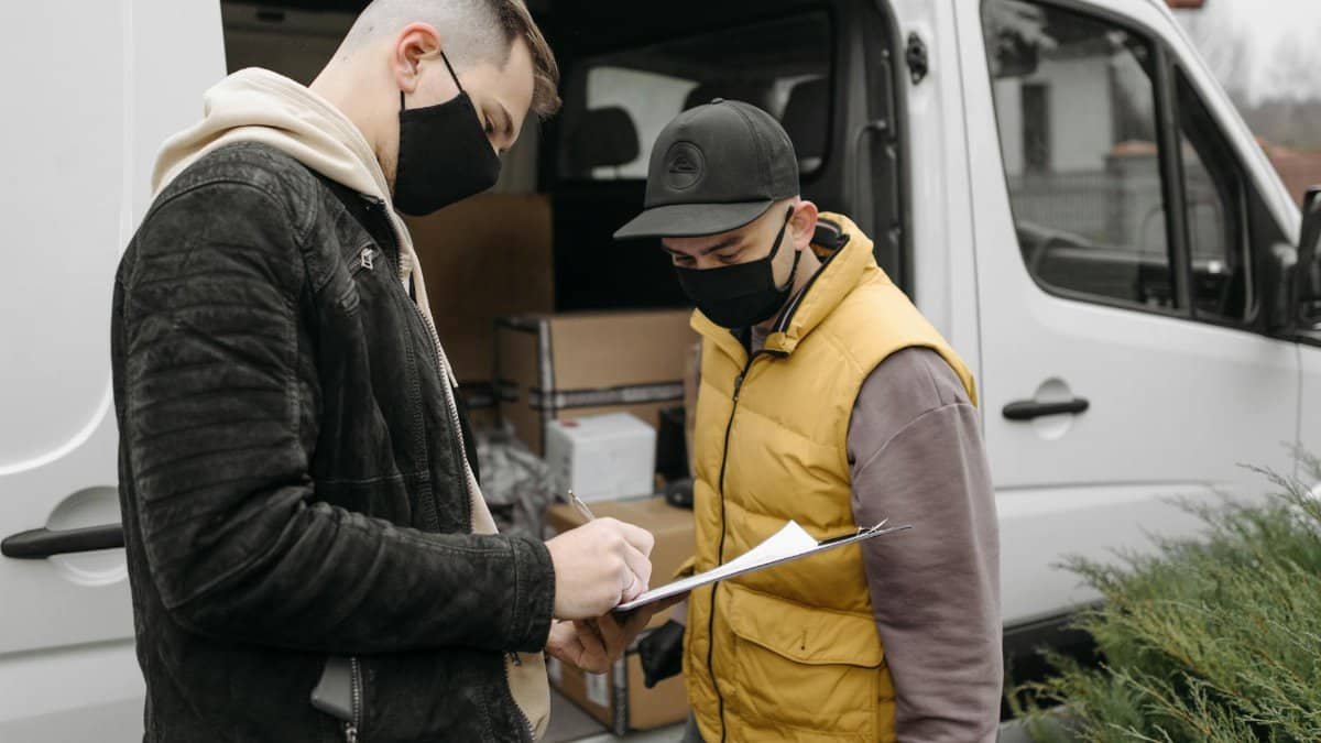 Masked individuals sign paperwork for a courier delivery beside a van. Safe, efficient logistics.