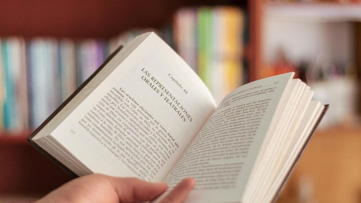 A hand holds an open book with blurred bookshelves in the background.