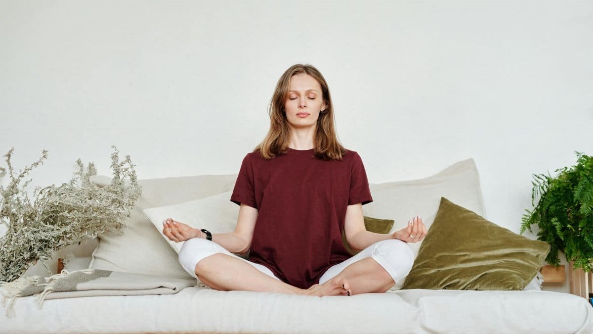Woman meditating indoors on a sofa, exuding calmness and mindfulness, surrounded by greenery.