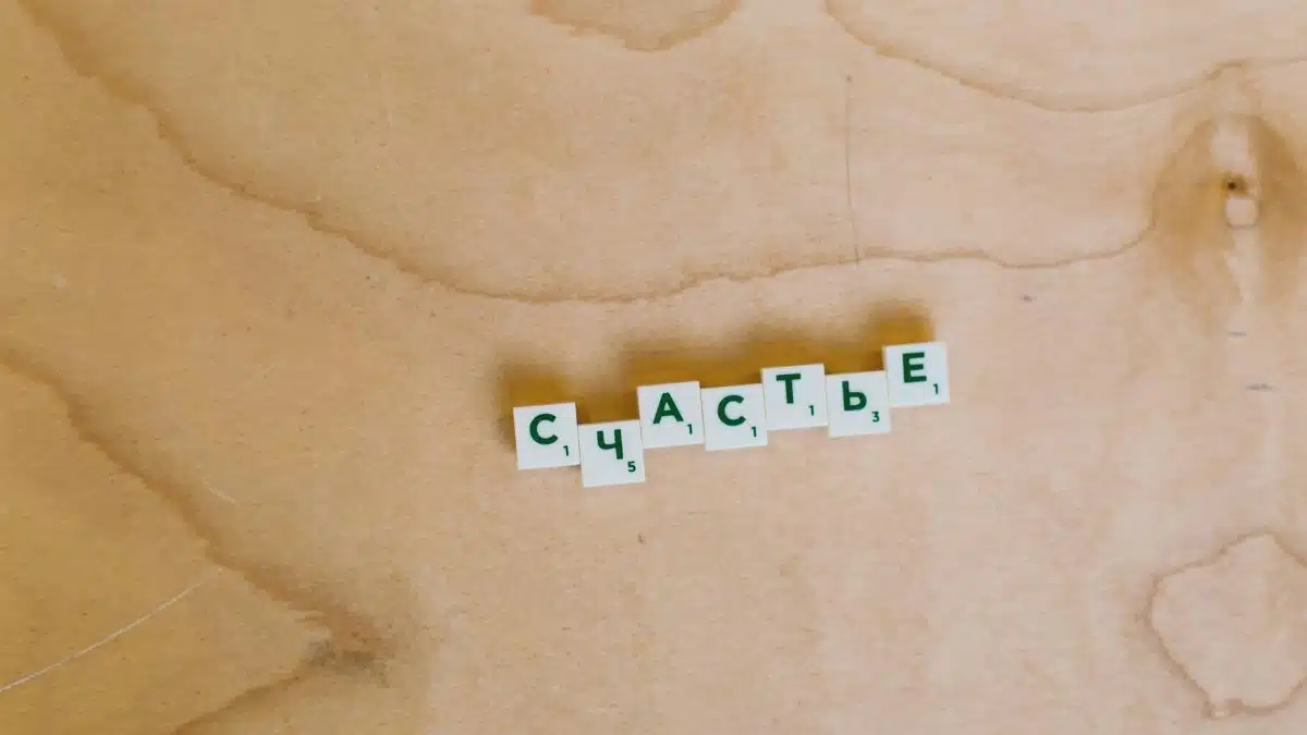Scrabble tiles spelling 'Happiness' in Cyrillic on a light wooden surface.