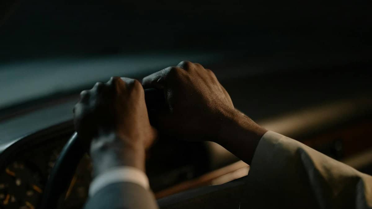A driver's hands grasp the steering wheel in a dimly-lit vehicle interior, conveying focus and control.