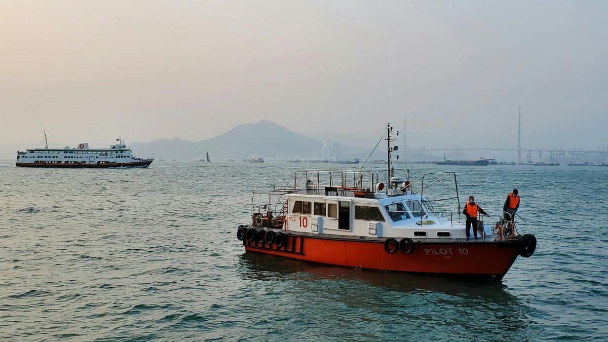 A pilot boat operating near a city skyline, with crew members on deck. Seascape under a clear sky.