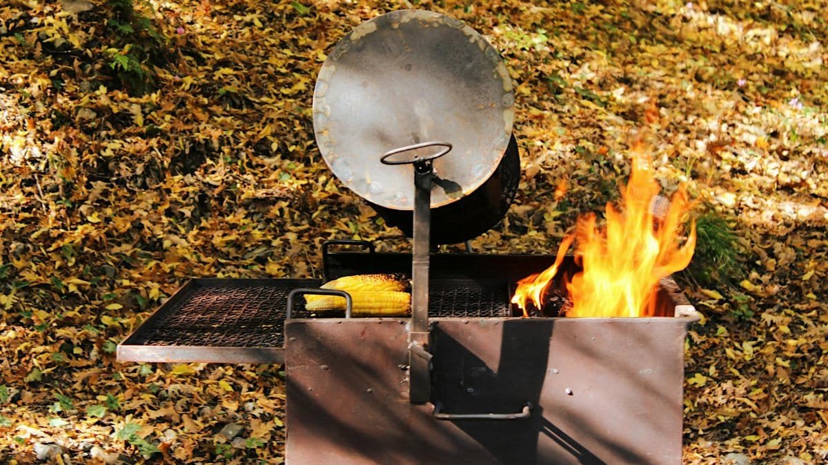 Rustic grill with fire and corn in autumn leaves setting, perfect for outdoor cooking vibes.