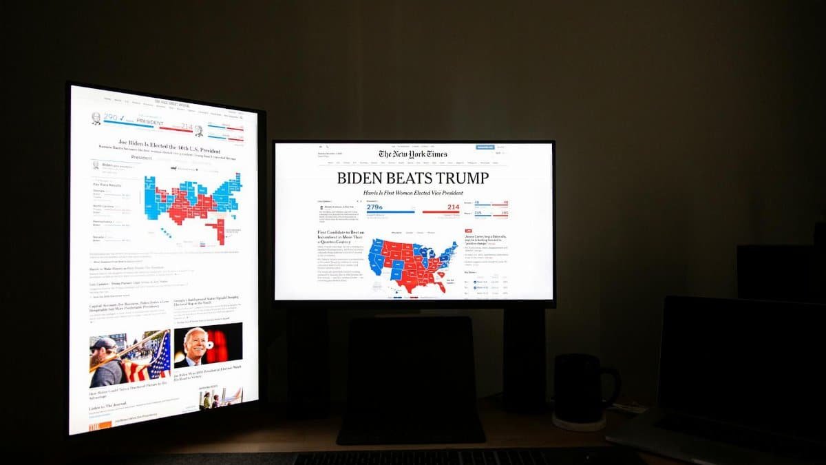 Dual computer screens in a dark room display election results indicating Biden's victory over Trump.