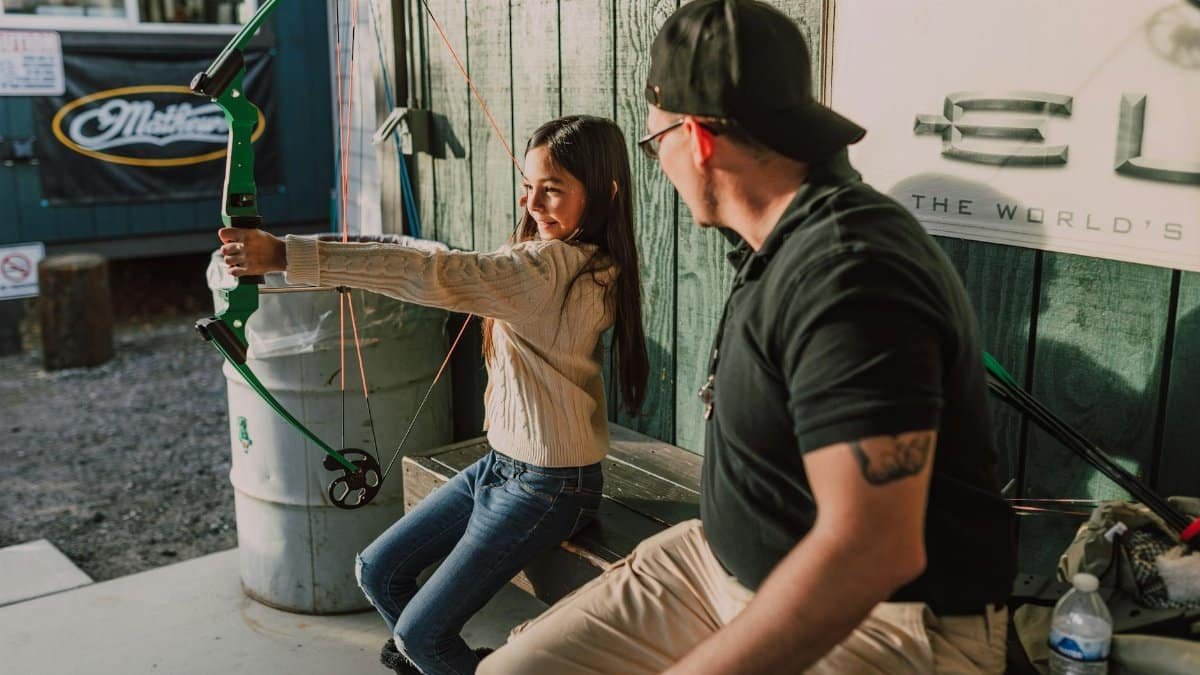 A young girl practices archery with a mentor outdoors, holding a compound bow.