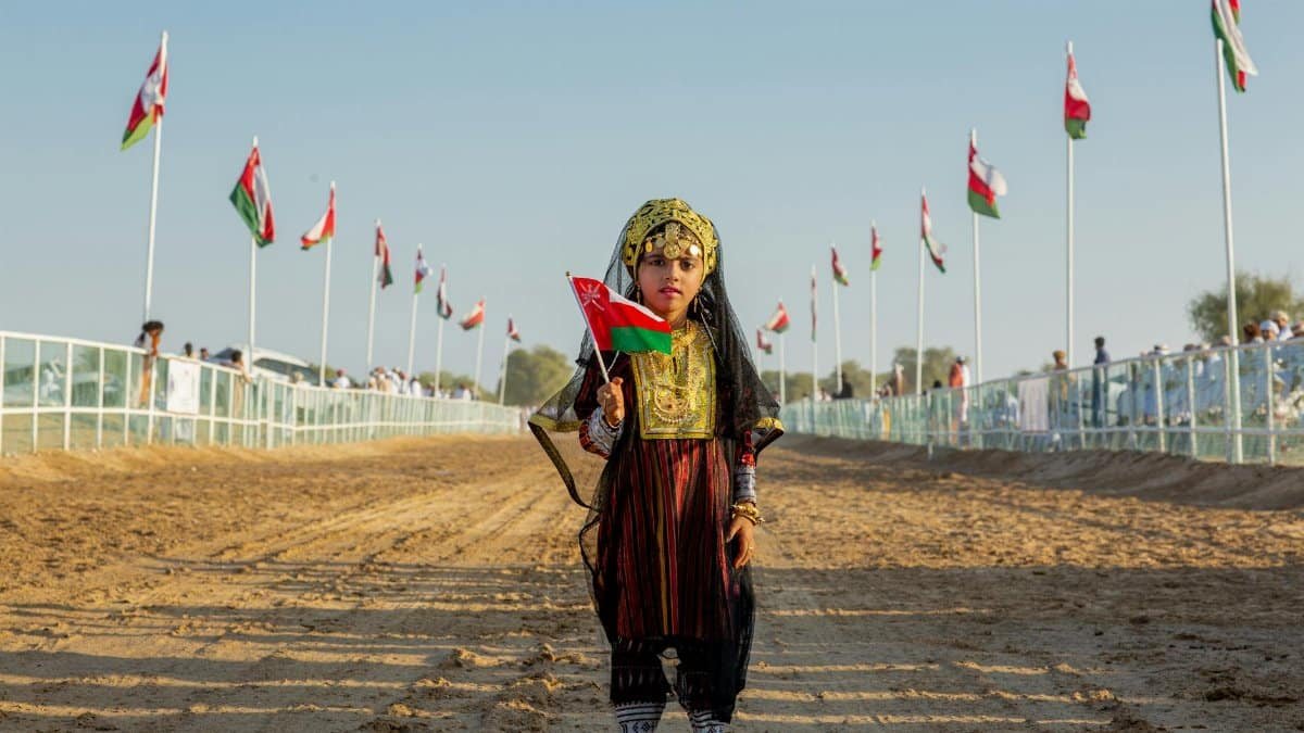 A young girl in traditional Omani clothing holds the flag of Oman during a festive outdoor celebration.