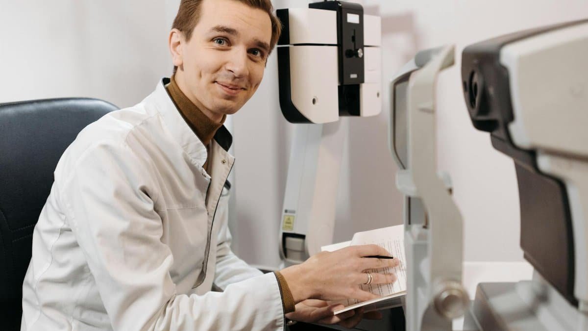 Smiling optometrist wearing a lab coat, taking notes beside eye examination equipment in a clinic.