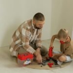 A father and son bonding over a building project with toy tools on a rug indoors.