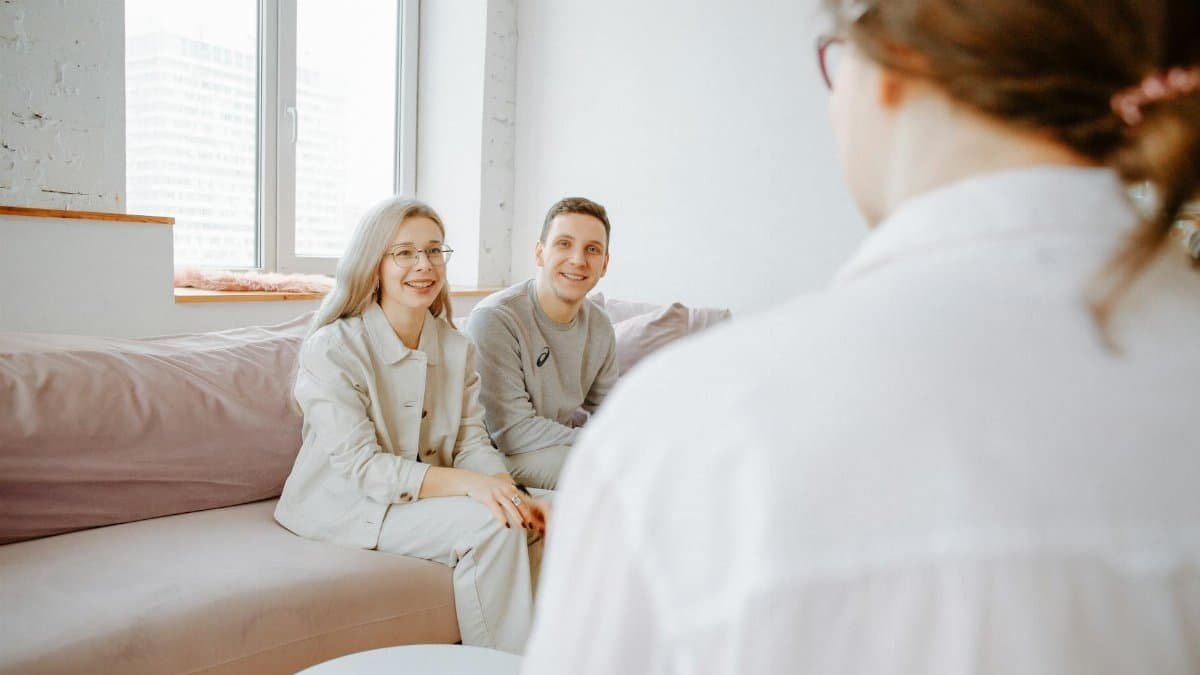 A couple smiles while sitting on a sofa during a counseling session indoors.