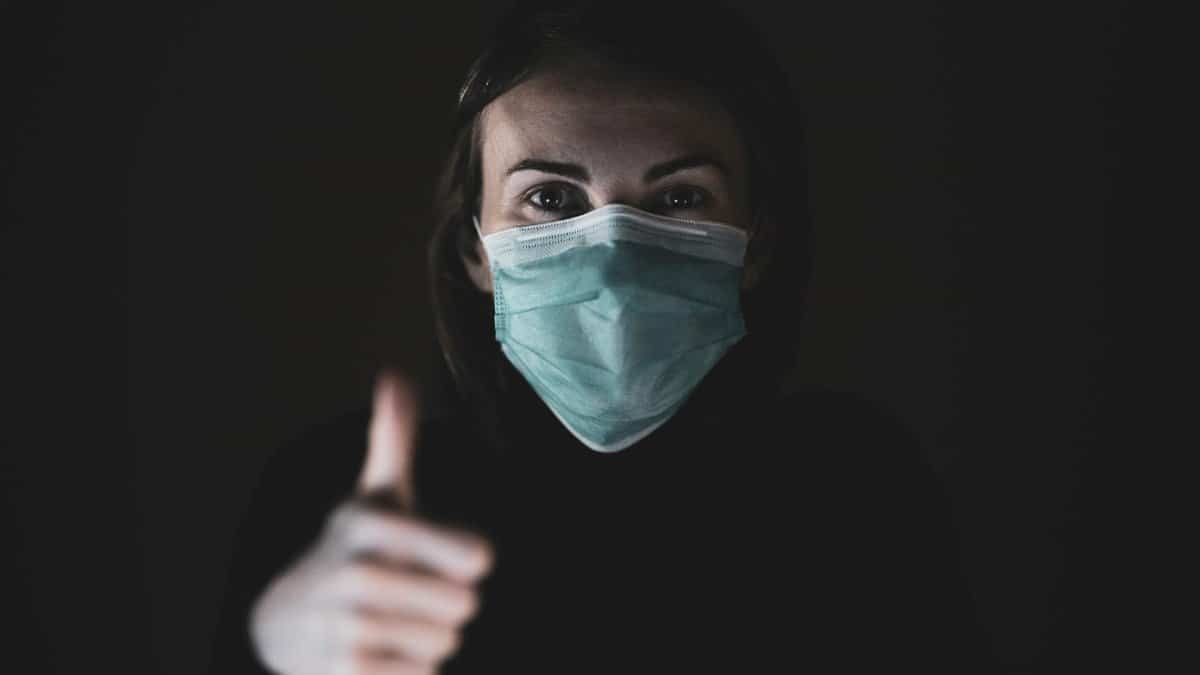 A woman wearing a mask gives a thumbs up in a dark setting, promoting safety during the pandemic.