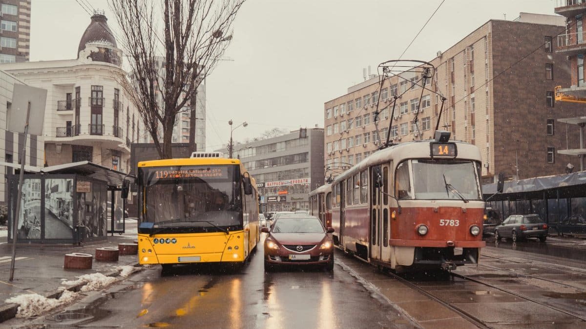 A snowy city street in Kyiv with a tram, bus, and car navigating through winter weather.