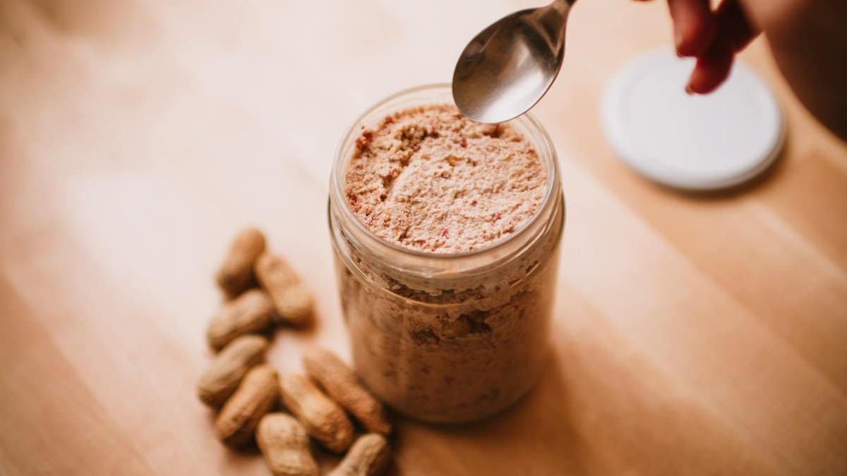 A glass jar of homemade peanut butter on a wooden table with peanuts and a spoon.