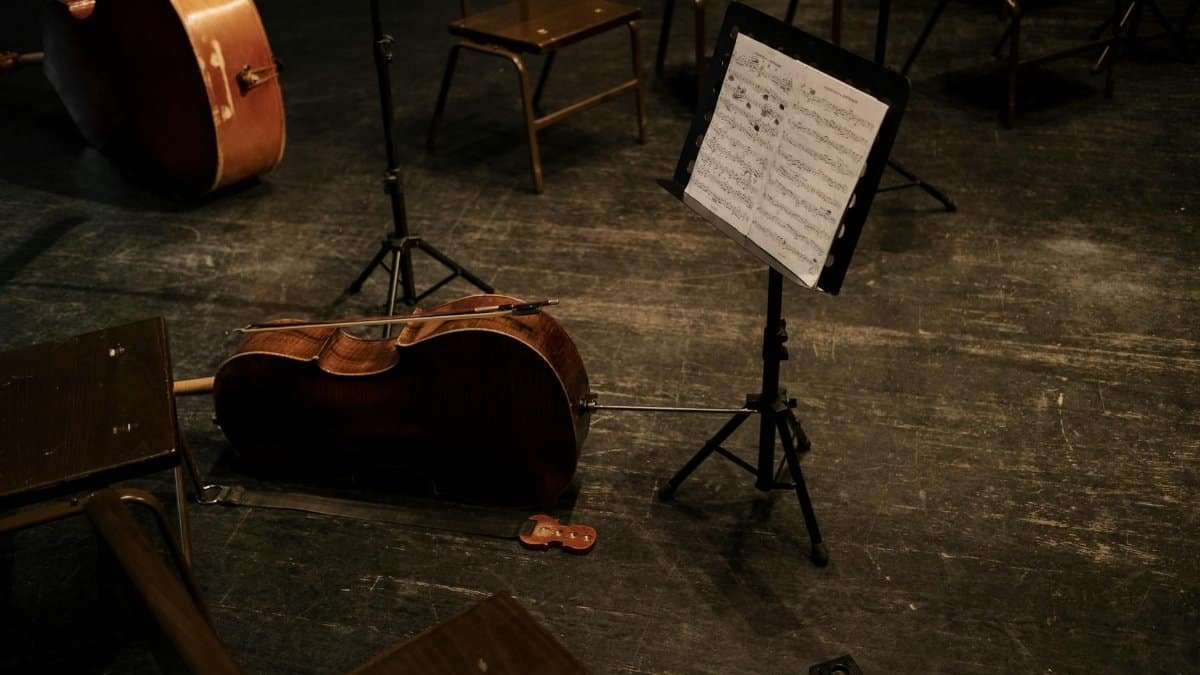 A cello rests on an empty stage with a music stand and scattered chairs, evoking classical music ambiance.
