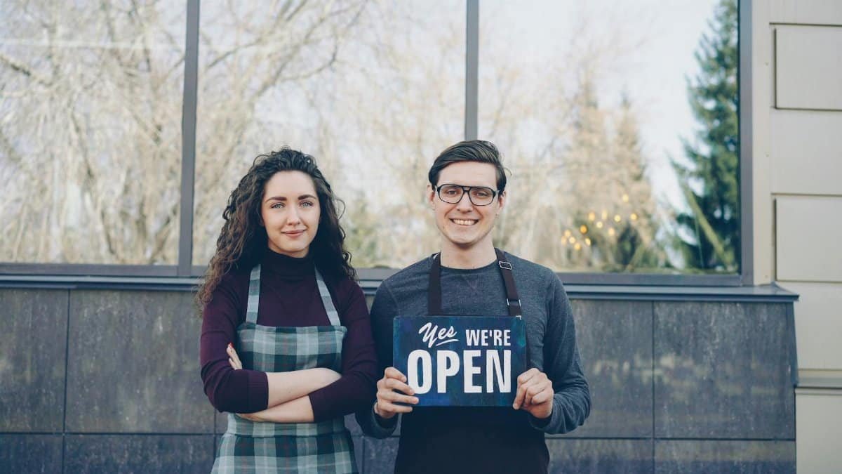 Two business owners standing outside, holding a 'Yes We're Open' sign, welcoming customers.
