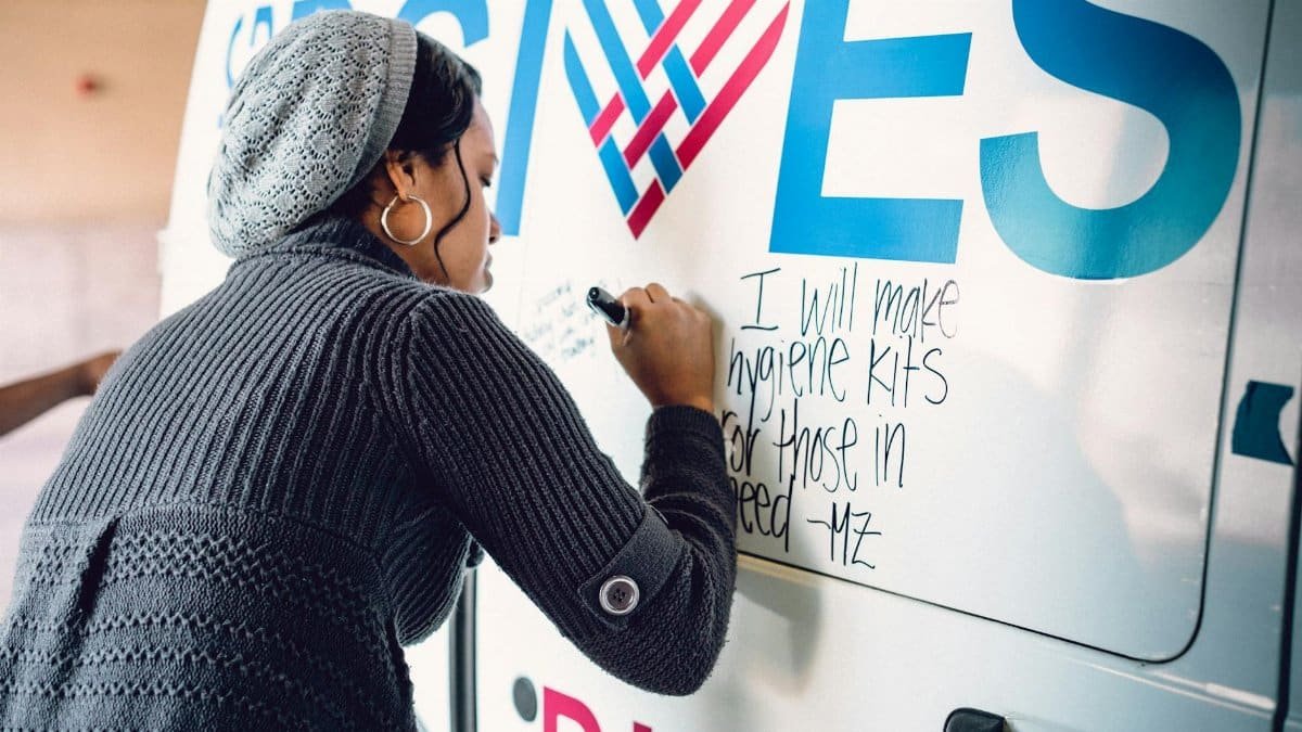 A black woman writes an inspirational note on a charity van, participating in a community event.