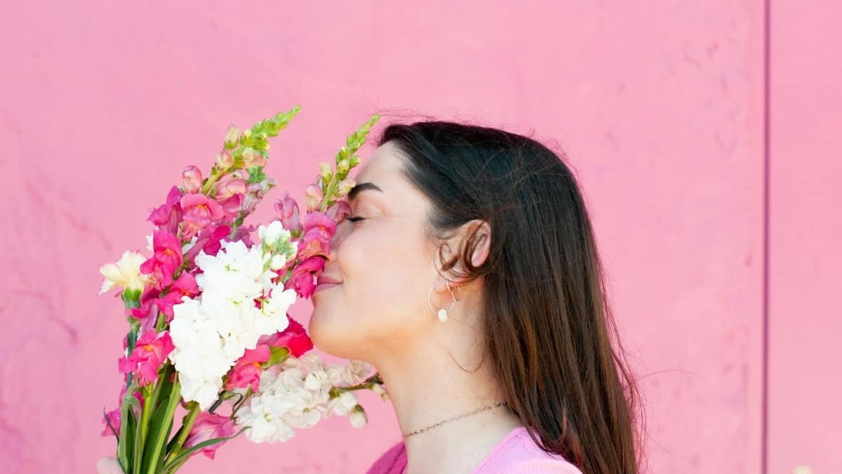 A woman with long brown hair enjoys the scent of a vibrant bouquet of flowers against a pink backdrop.