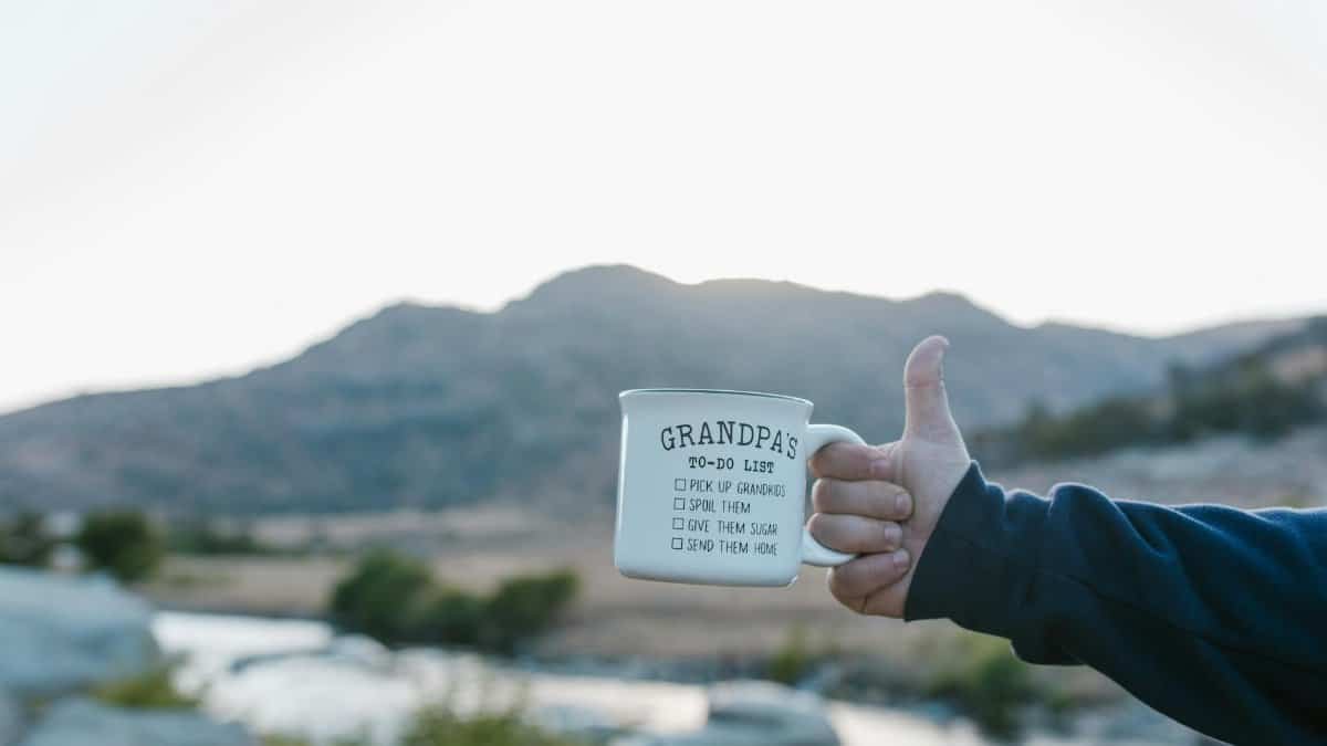 Outdoor close-up of a ceramic mug with text held up against a scenic mountain backdrop.