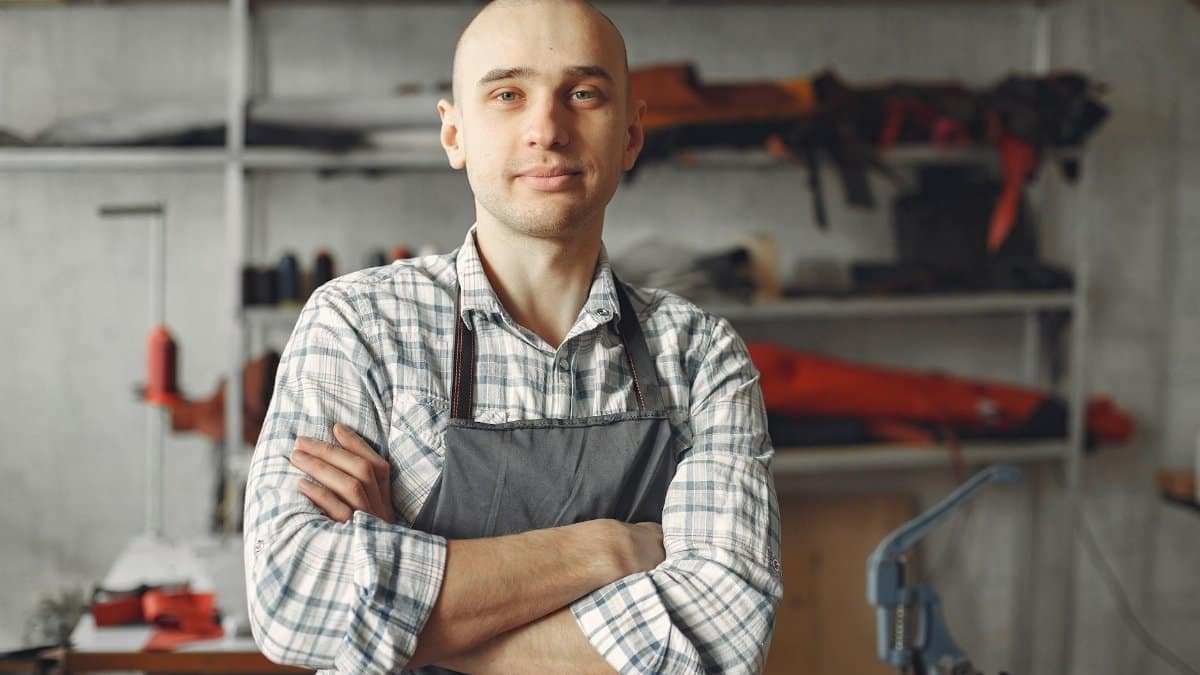 Positive young artisan in check shirt and apron standing with arms folded and looking at camera in contemporary workshop in daylight