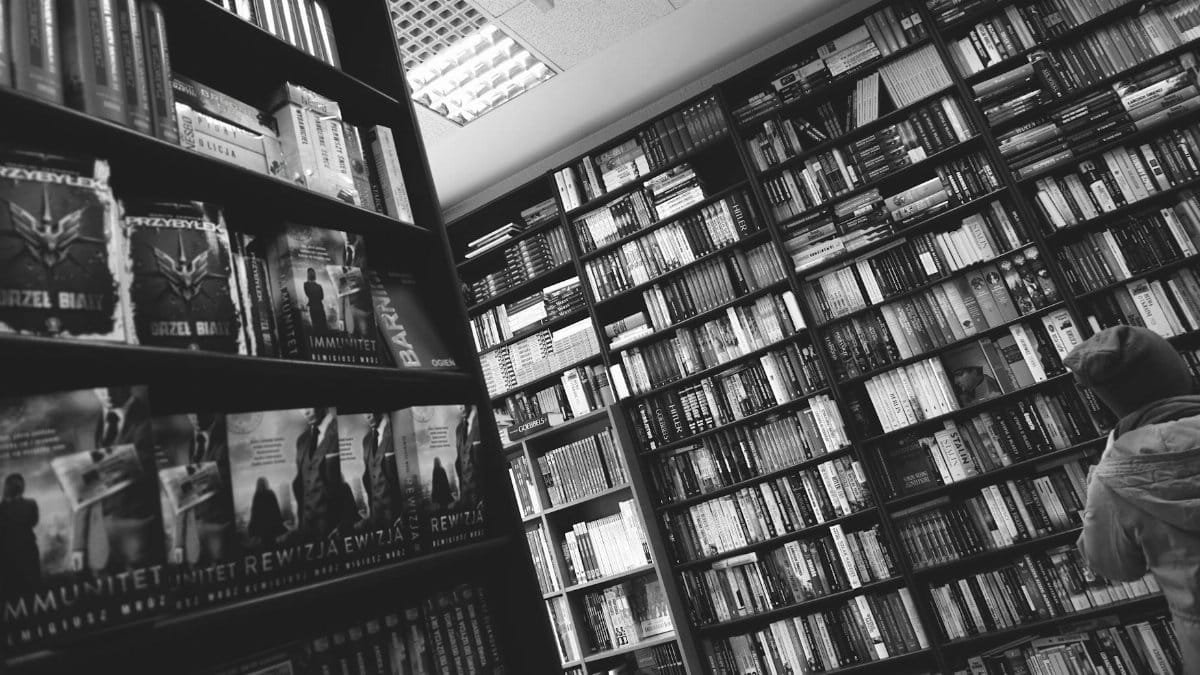 Black and white image of a person browsing bookshelves in a library or bookstore.