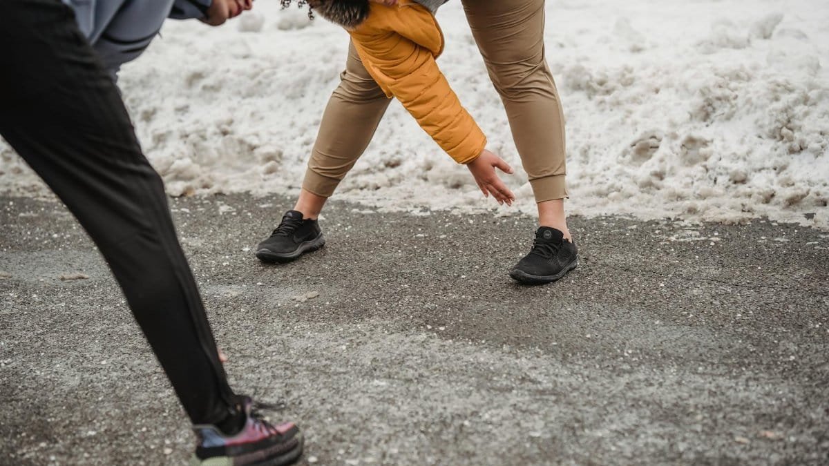 Active African American overweight female with black personal coach doing warm up exercises while bending forward on sidewalk during workout in snowy terrain