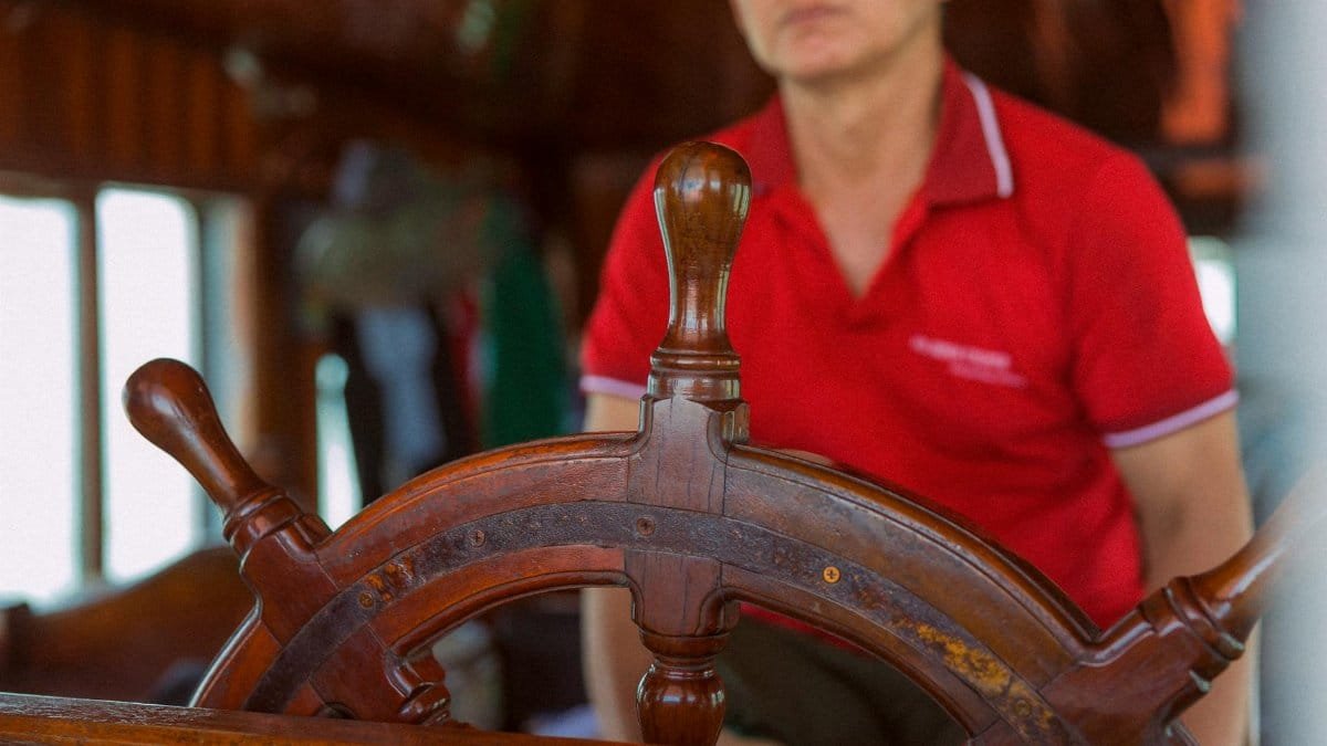 Close-up of a man in red shirt at a ship's wooden helm, steering indoors.