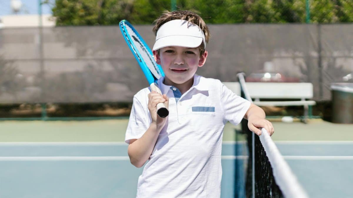 A smiling young boy stands at a tennis court net holding a racket, ready for play.