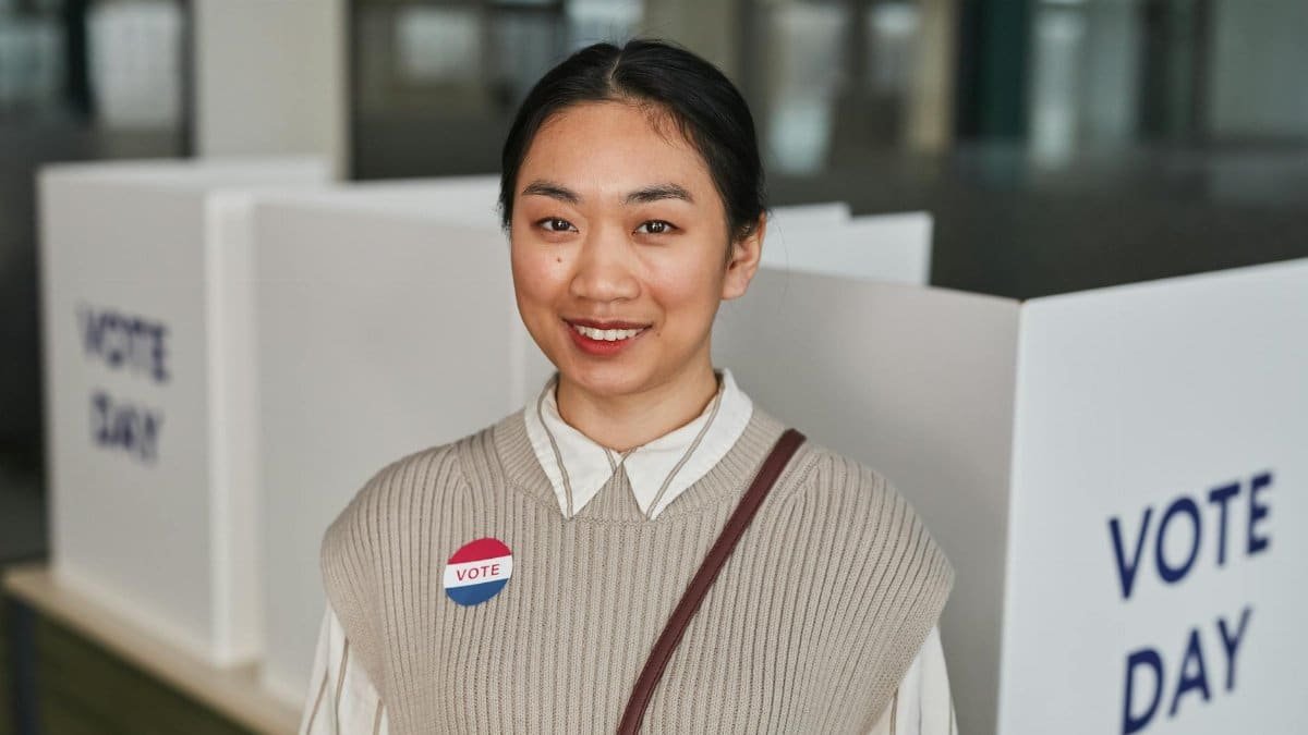 Asian woman smiling at polling station with 'Vote' badge on Voting Day.