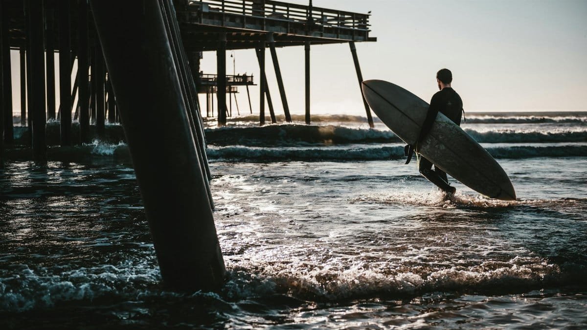 A surfer walks with a surfboard by the pier at Pismo Beach, California, highlighting a serene sunset scene.