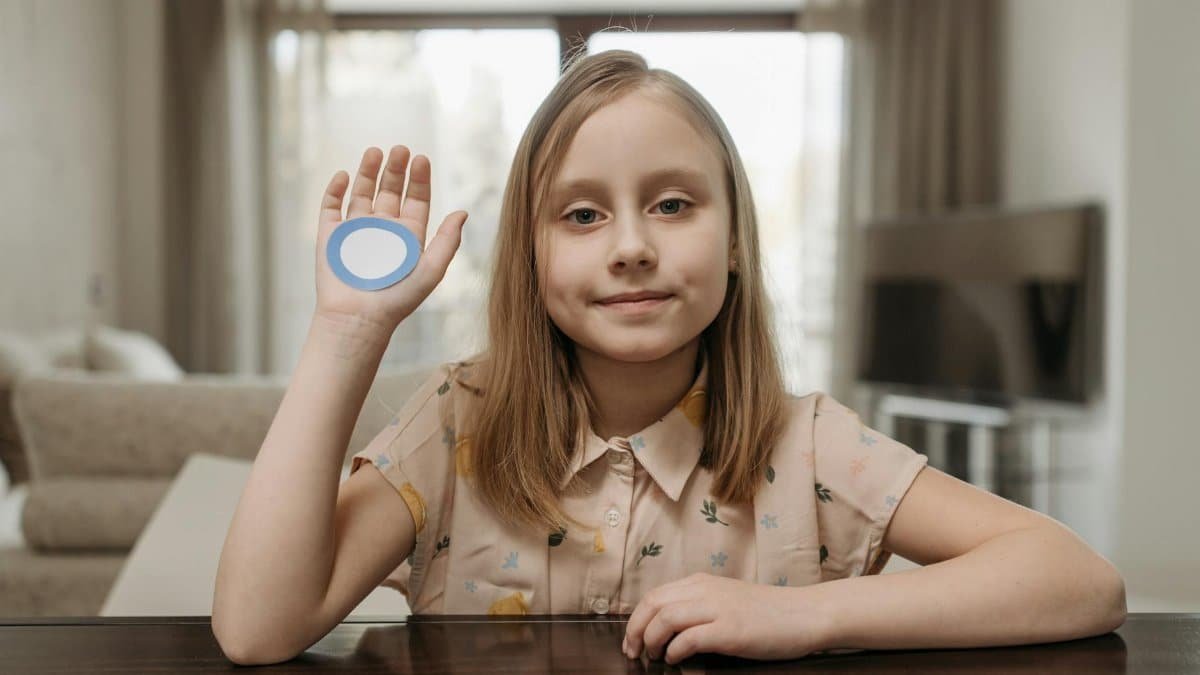 Young girl sitting indoors, raising hand with blue diabetes awareness circle symbol.
