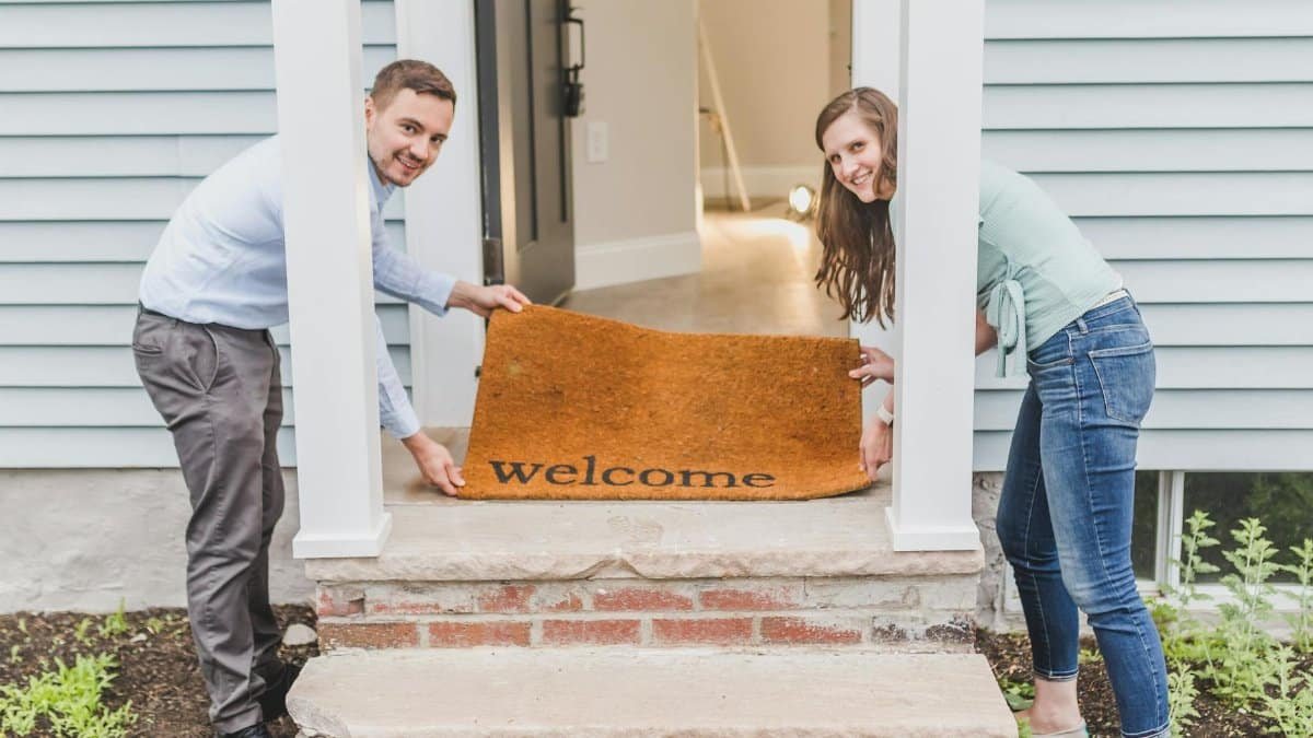 Couple smiling while holding a 'welcome' mat at their home's front door, symbolizing a warm greeting.