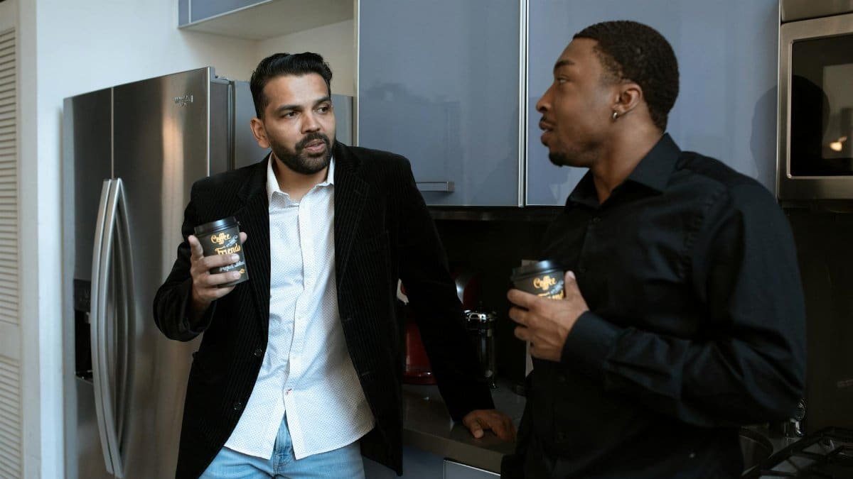 Two men having a casual discussion while enjoying coffee in an office kitchen setting.