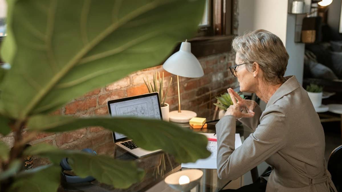 Senior woman in a modern home office focusing on work at a laptop next to a brick wall.