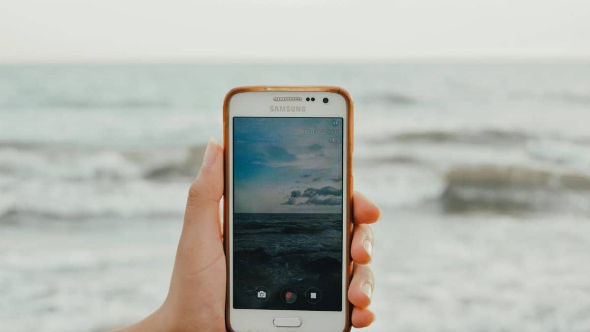 A hand holds a smartphone capturing a picturesque view of the ocean waves at the beach.