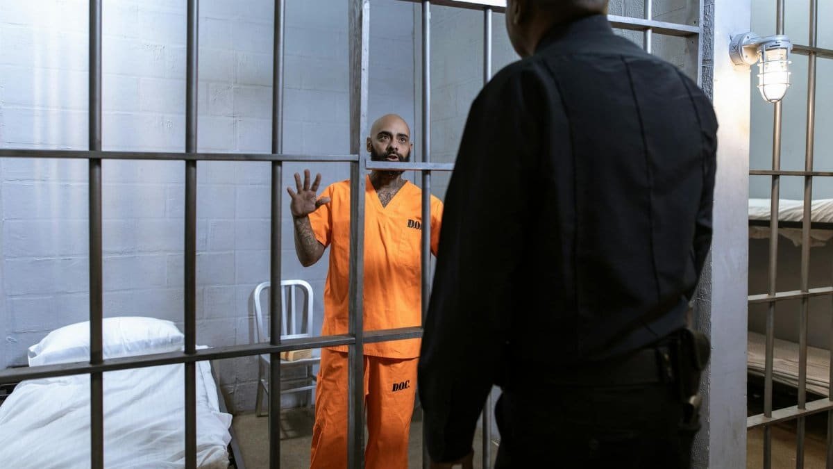 A male prisoner in an orange uniform stands behind bars facing a guard in a jail cell.
