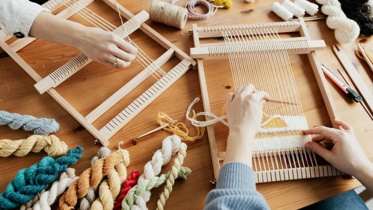 Two people weaving on wooden looms with colorful yarns in a workshop setting.