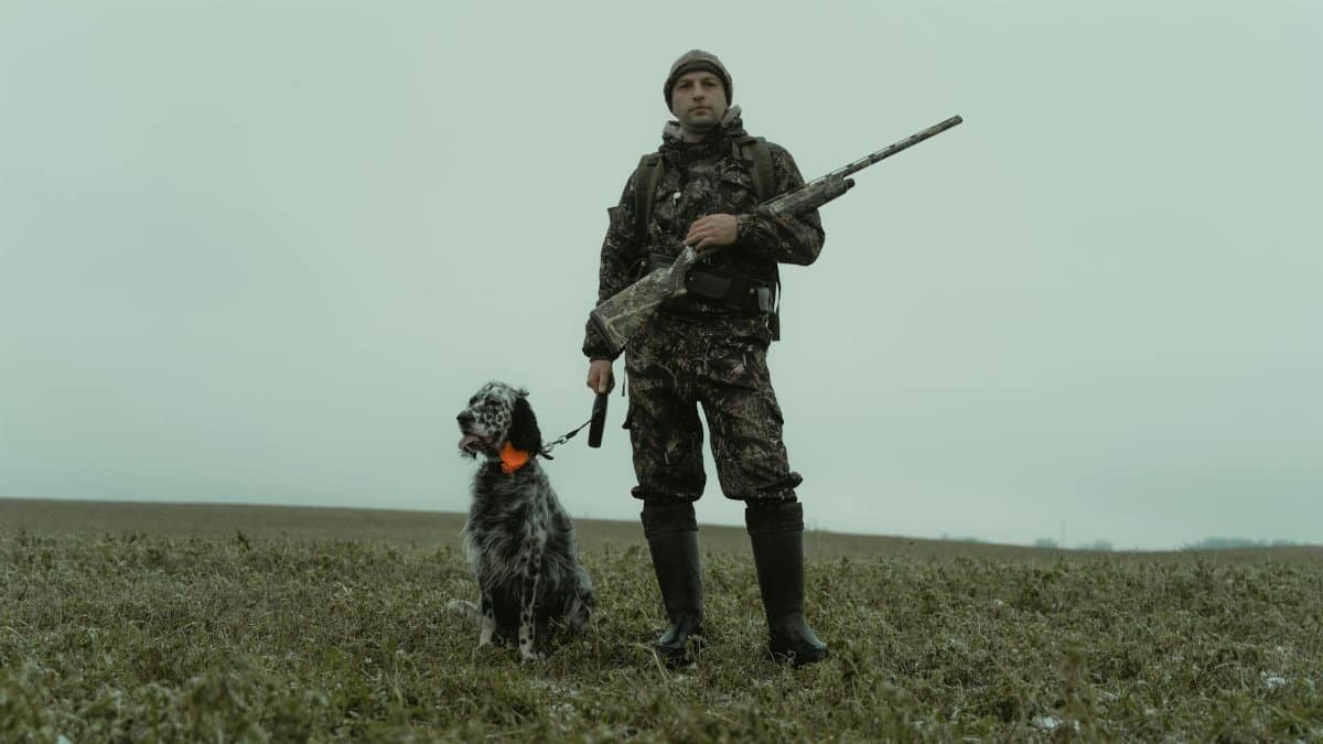 Man in camouflage with hunting dog and rifle in winter field.