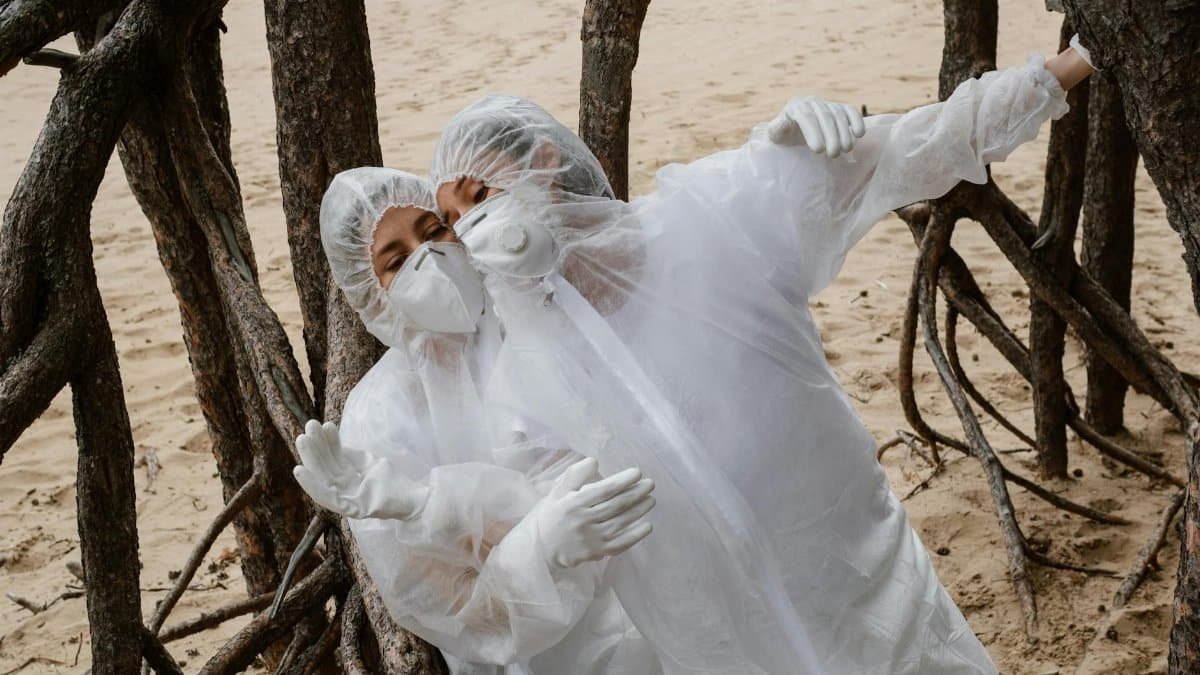 Two individuals in protective clothing and masks pose among tree roots on a sandy beach.