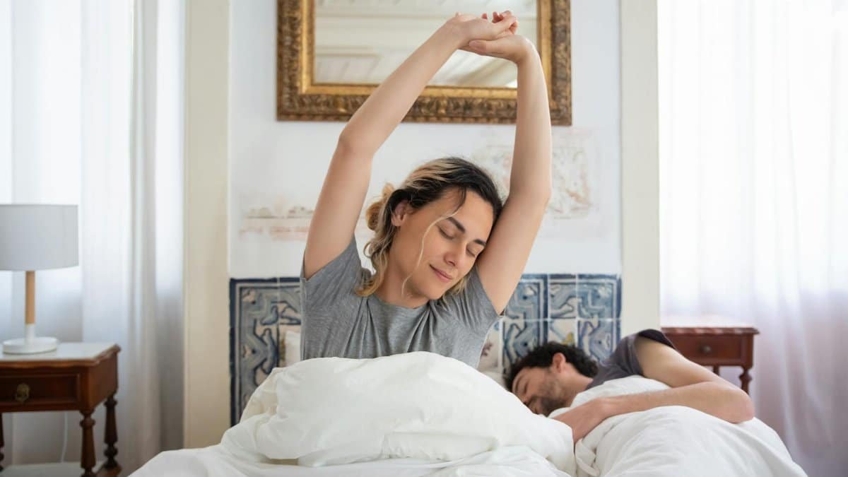A couple waking up peacefully, woman stretching in bed in a cozy bedroom setting.