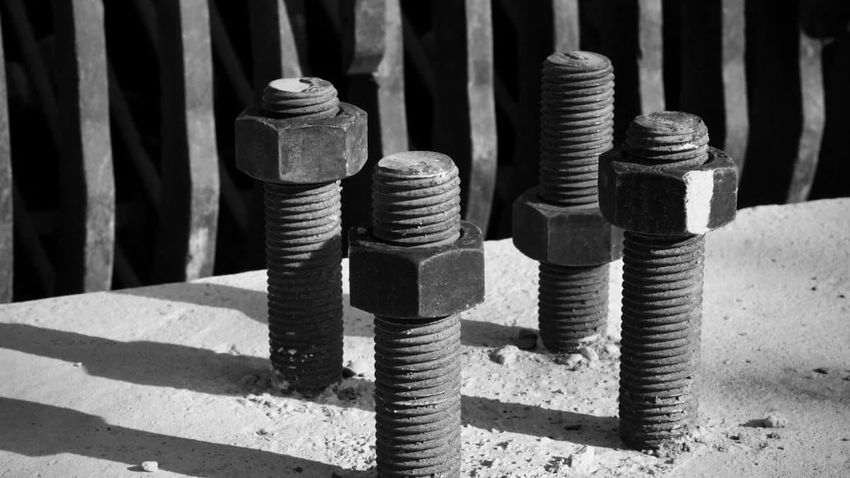 Black and white image of four metal bolts on a concrete surface with shadows, highlighting industrial theme.