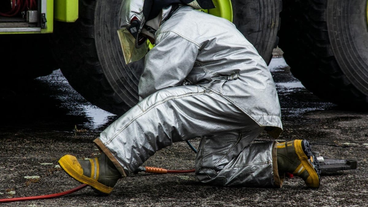 Firefighter in silver protective gear kneeling by a vehicle, ensuring safety measures.