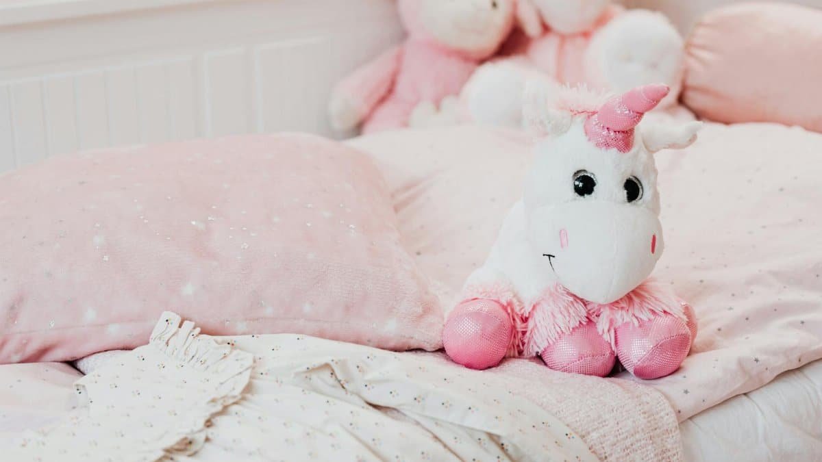 A soft unicorn plush toy sits on a pastel pink bed with pillows and blankets in a child's room.