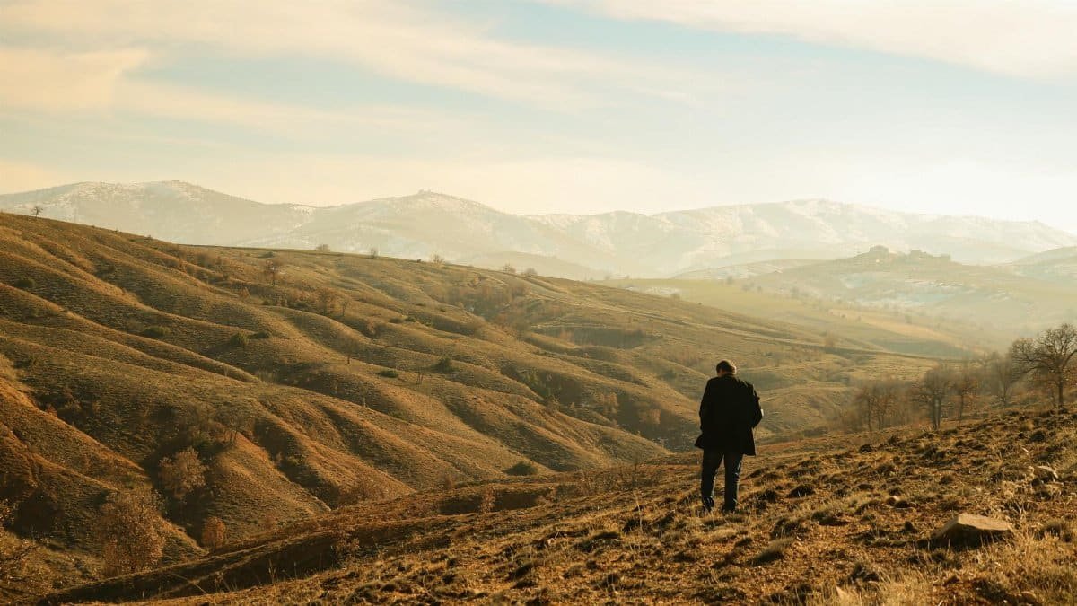 A lone person standing amidst rolling hills and distant mountains under a clear sky.