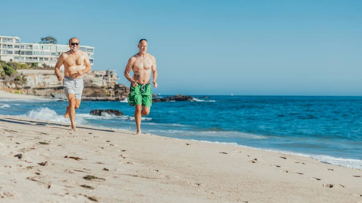 Two men jogging shirtless on a sunny beach by blue ocean waves.