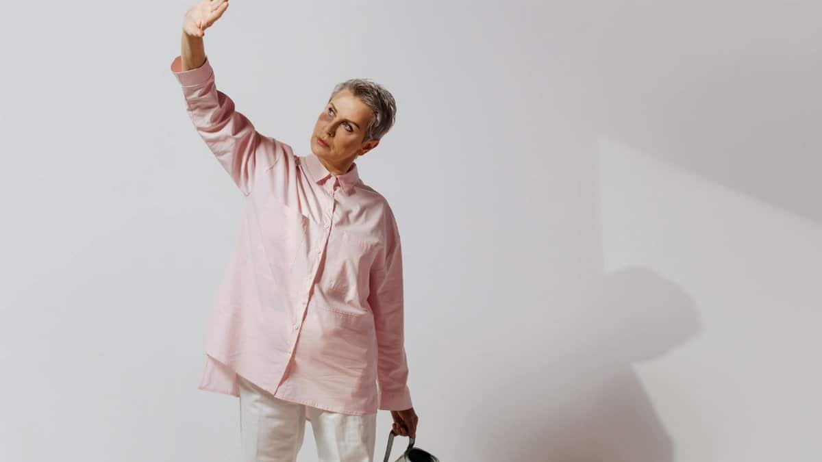 Elegant senior woman in pink shirt holding a watering can, gesturing gracefully in a minimalist setting.