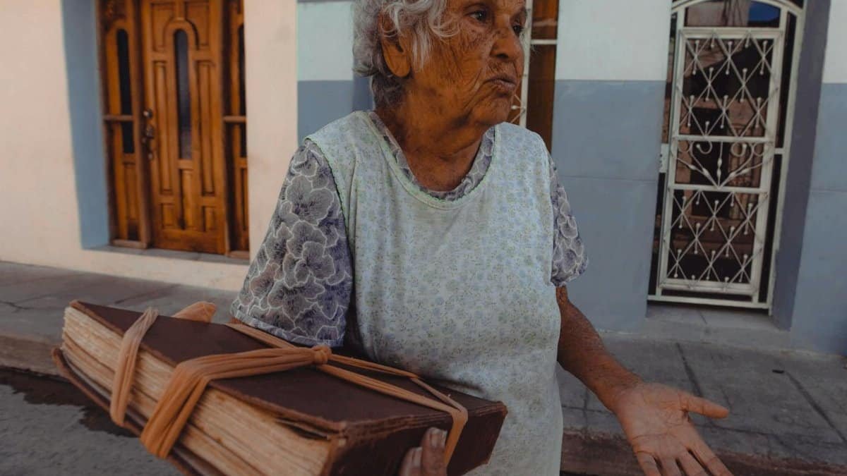 An elderly woman stands on the street holding a large, historic book, conveying wisdom and storytelling.