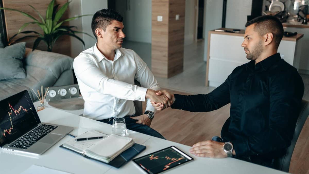 Two businessmen shake hands over financial charts and gadgets, symbolizing a successful deal.